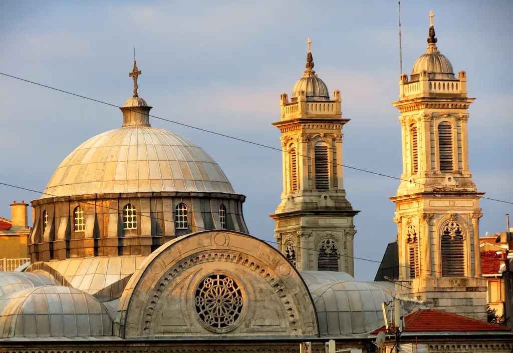Akşam ışığında Aya Triada Kilisesi; altın renkli kubbe ve kasnak pencereleri, iki çan kulesinin üst katları ve önde gül pencere.