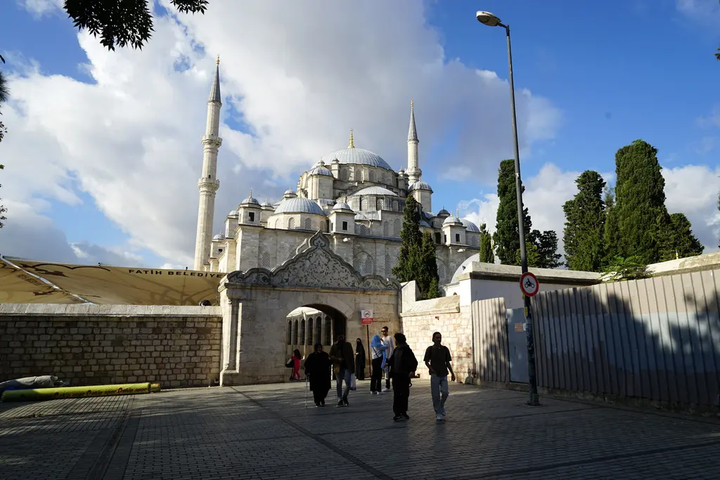 Fatih Külliyesi'nin Çorbacı Kapısı; avlu duvarında sivri alınlıklı taş kemerli kapı, içinden geçen insanlar, arkada gri kubbeler, iki minare ve serviler.