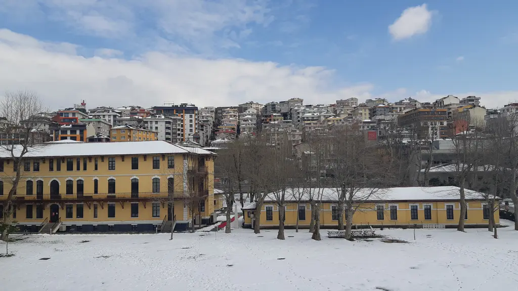 Santralistanbul'da karlı yerleşke; kar kaplı çimenler, yapraksız ağaçlar, sarı cepheli uzun tarihi binalar ve arkada yamaçtaki evler.