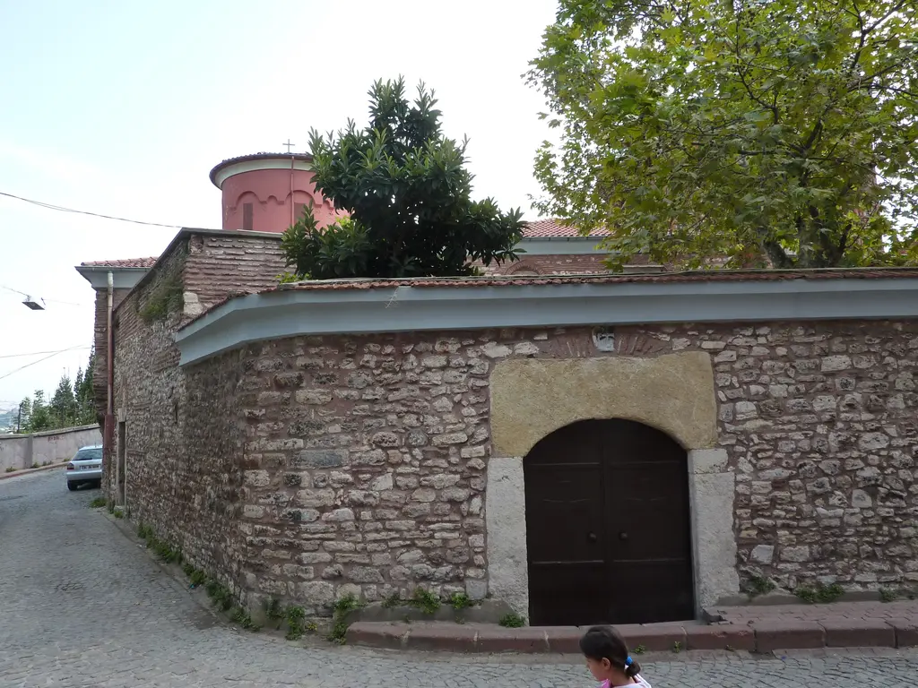 Kanlı Kilise'nin çevre duvarı; köşeden dönen moloz taş duvar, ortada taş söveli kemerli ahşap kapı, arkada ağaçlar ve kırmızı kubbe.