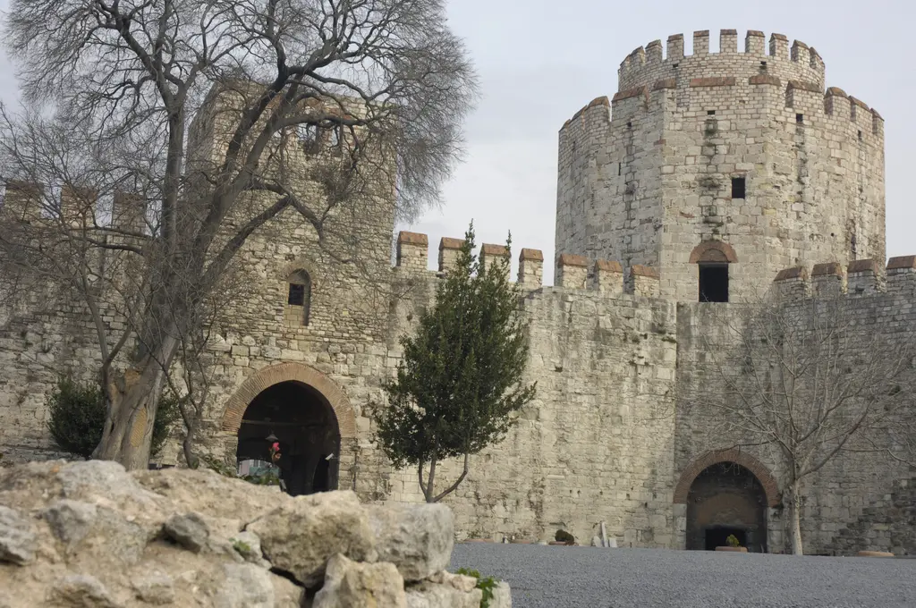 Yedikule Hisarı'nın giriş kapısı; ortada tuğla örgülü sivri kemer, iki yanda kule gövdeleri, sağda mazgallı büyük yuvarlak kule, önde yapraksız ağaçlar ve dağınık taş bloklar.