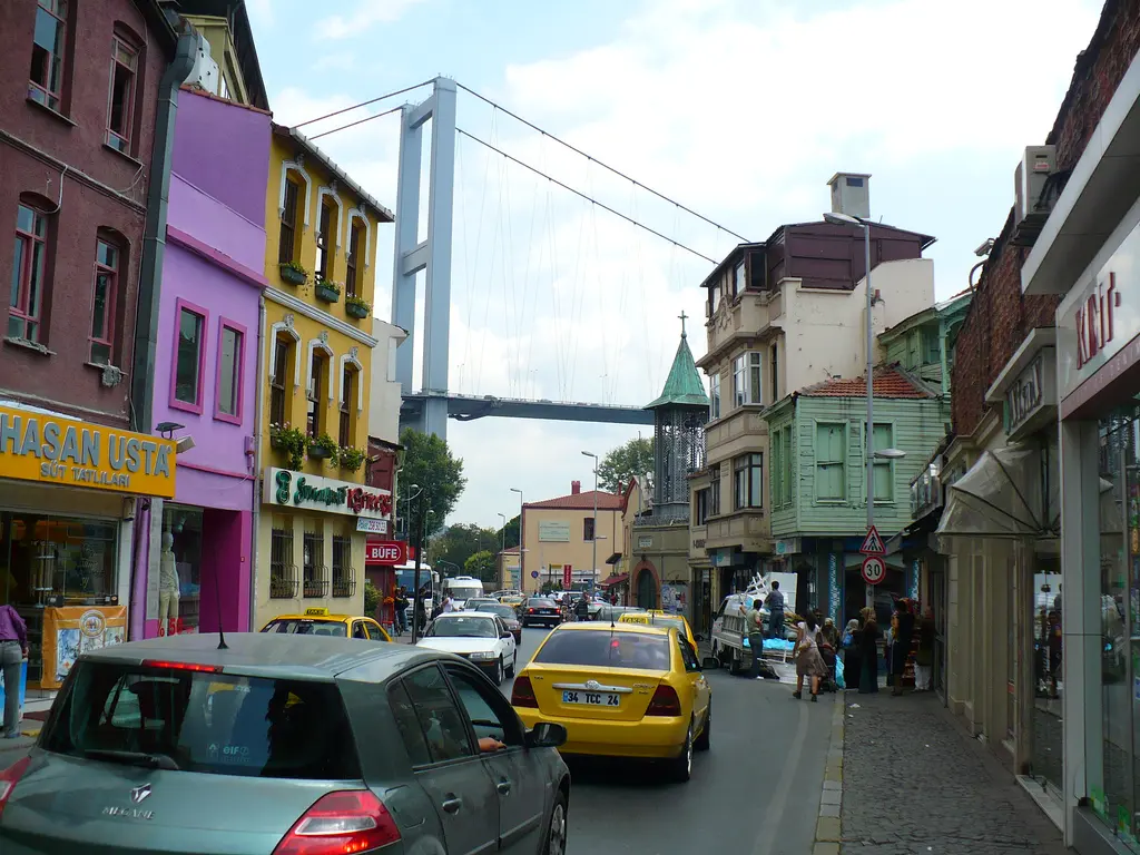 Ortaköy'de cadde boyunca mor, sarı ve kırmızı cepheli evler, sarı taksiler, arkada köprü ve çan kulesi.