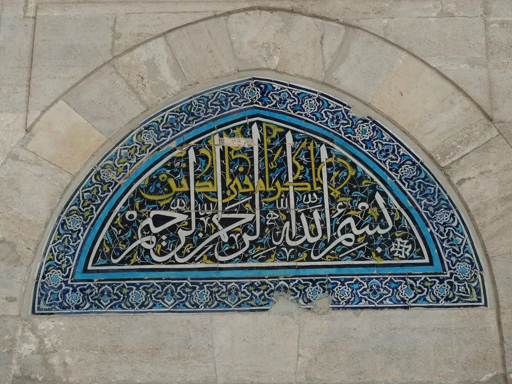 Fatih Camii avlusunda yarım daire çini alınlık; koyu kobalt zemin üzerinde beyaz sülüs hat, turkuaz ile sarı rumi kıvrımlar ve mavi beyaz palmet bordür.
