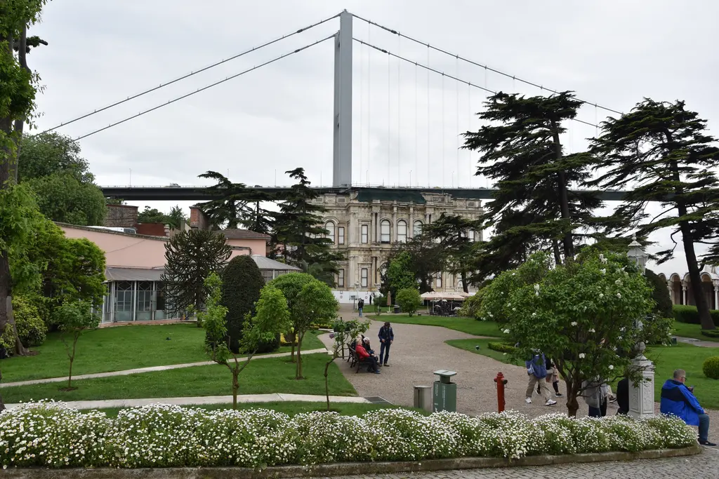 Beylerbeyi Sarayı'nın bahçesi; çimenler, çiçek tarhları, büyük ağaçlar, gezinti yolu ve arkada köprünün kulesi.