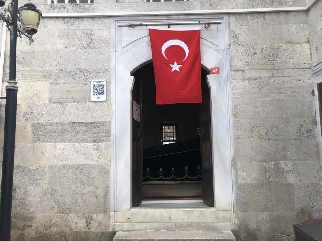Şemsi Ahmed Paşa Türbesi'nin girişi; kesme taş cephe, kemerli kapı, kapıya asılı Türk bayrağı ve fener.