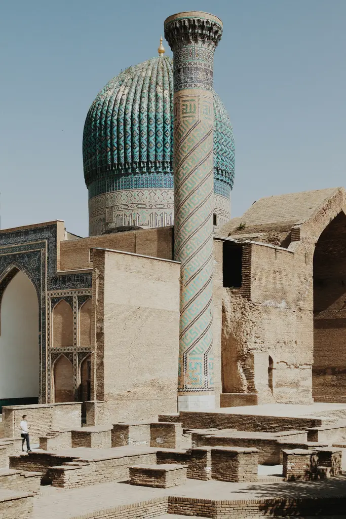 Semerkant'ta Gur-e Amir'in kaburgalı turkuaz kubbesi ve spiral çinili köşe minaresi; solda çinili portal, önde kazıyla açığa çıkan tuğla temeller ve ölçek veren bir kişi.