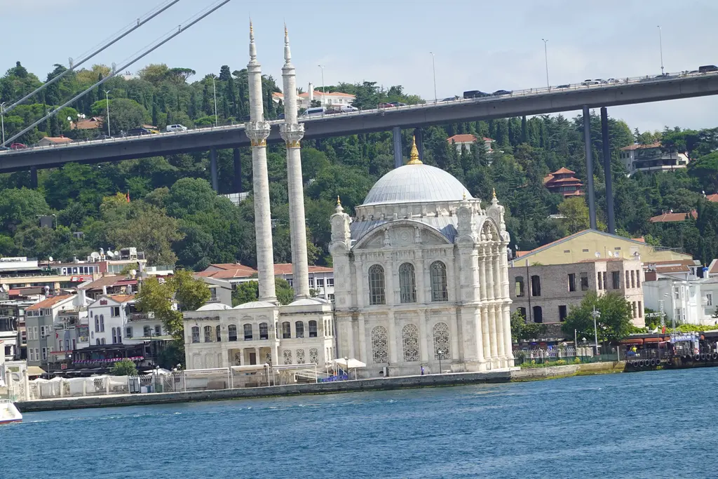 Denizden Ortaköy Camii; Boğaz kıyısında beyaz kubbeli cami ve minareler, arkada köprünün ayağı, yamaçtaki evler ve ağaçlar.