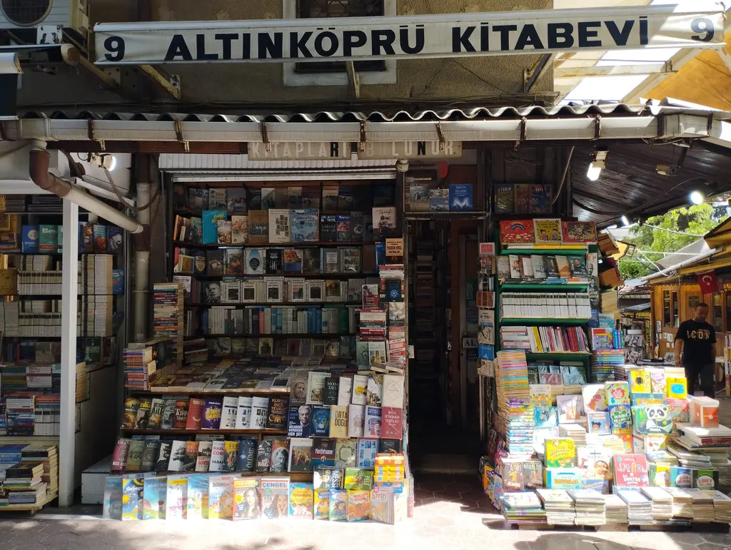 Sahaflar Çarşısı'nda büyük beyaz tabelalı bir kitabevi; metal saçak, iki yanda döner kitap standları, yere istiflenmiş kitaplar ve sağda avluya bakan gölgeli koridor.