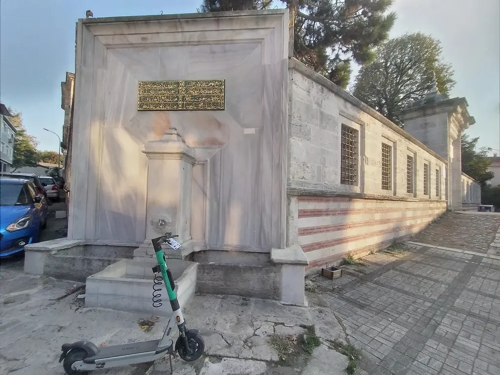 Ayazma Camii'nin köşe çeşmesi; dört köşe mermer gövde, üstünde yaldızlı kitabe ve yanında uzanan çizgili avlu duvarı.