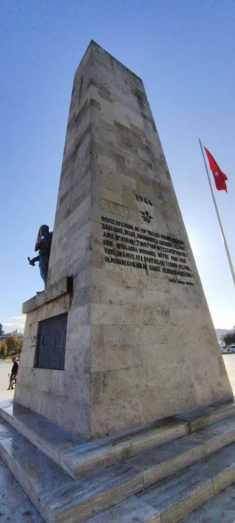 Barbaros Anıtı'nın arka yüzü; yüksek taş kütle üzerinde kazınmış dizeler, yanda bronz figür ve dalgalanan bayrak.