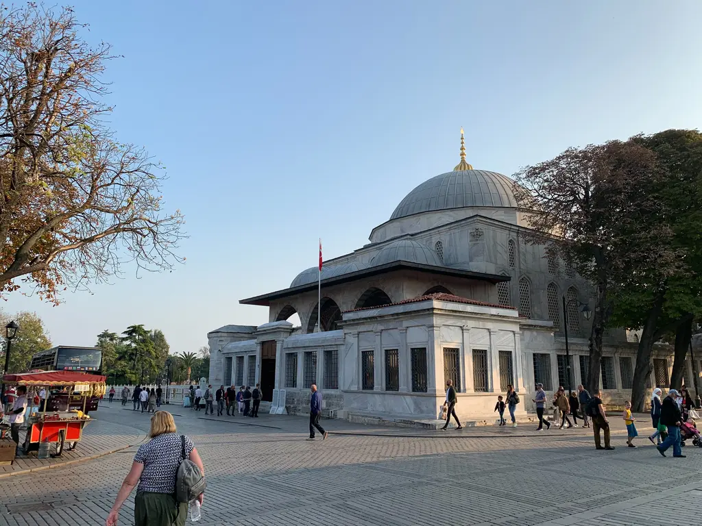 Meydandan Sultan Ahmed Türbesi ve revakı; kubbeli mermer türbe, önünde pencereli avlu duvarı, sonbahar ağaçları, simit arabası ve yayalar.