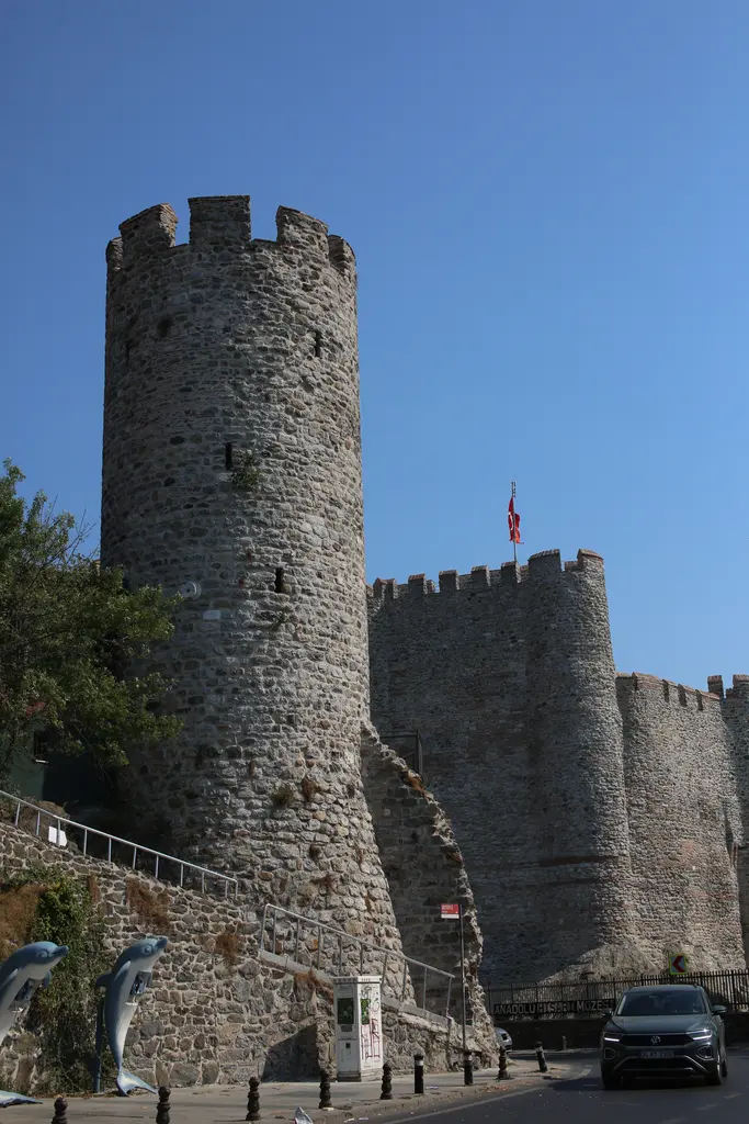 Anadolu Hisarı'nda kuleler; yüksek yuvarlak taş kule, arkasında mazgallı surlar ve önde yol.