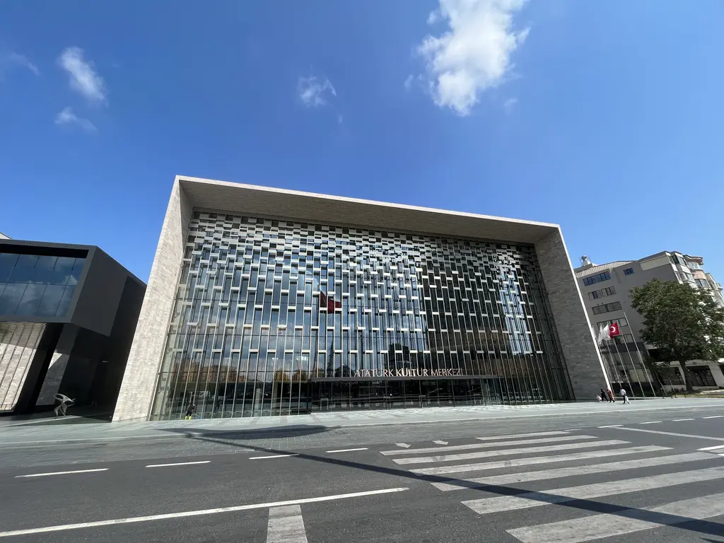 Atatürk Kültür Merkezi'nin meydana bakan cephesi; taş kaplı kalın çerçeve, içinde düzensiz desenli metal ve cam yüzey, önde cadde ve yaya geçidi.