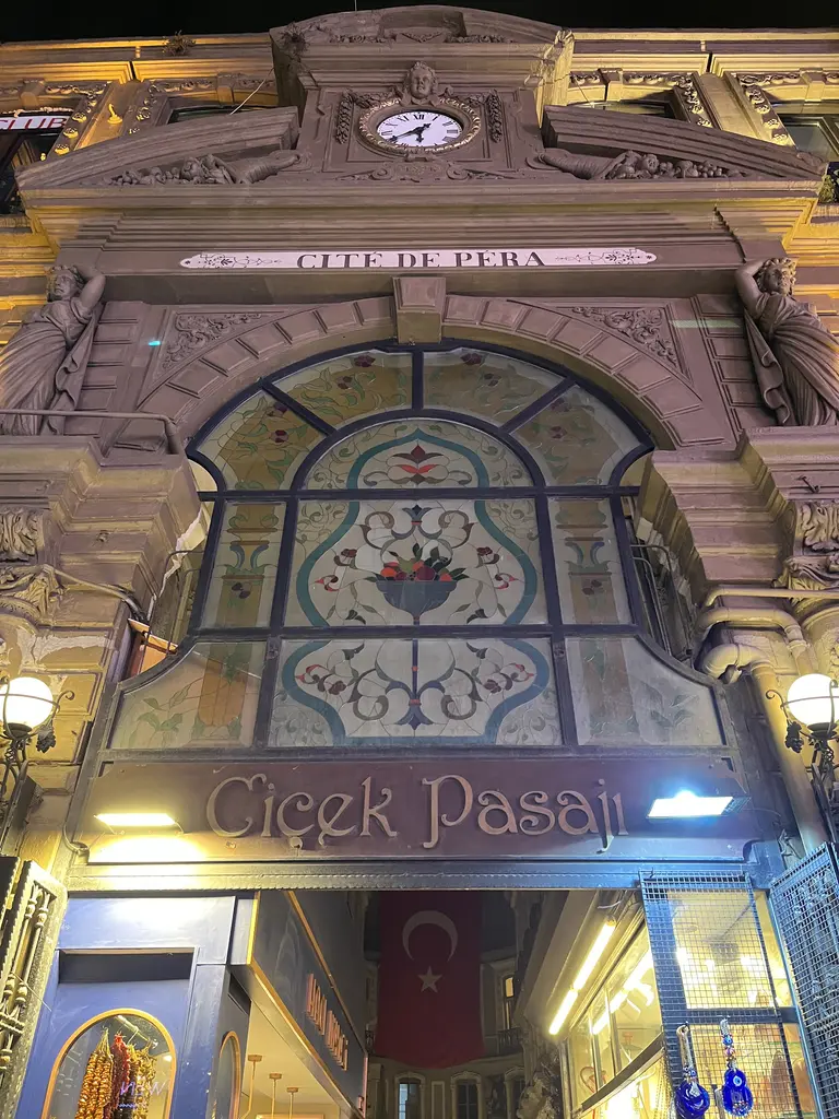 Gece Çiçek Pasajı'nın girişi; ışıklandırılmış renkli camlı kemer, "Çiçek Pasajı" tabelası, saat ve içeride asılı Türk bayrağı.