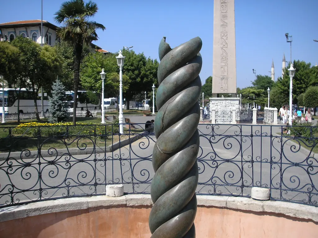 Önde Yılanlı Sütun'un burmalı üst kısmı, arkada Dikilitaş, palmiyeler, sokak lambaları ve parmaklıklı çukurun kenarı.