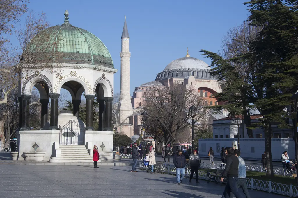 Alman Çeşmesi ile Ayasofya; önde yeşil kubbeli çeşme ve sütunlar, arkada Ayasofya'nın kubbesi ile minaresi, meydanda yürüyen insanlar.