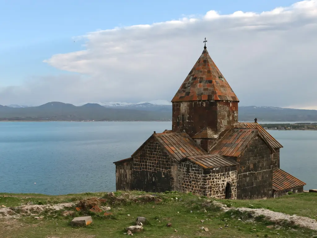 Ermenistan'da Sevan Gölü kıyısındaki tepede yükselen Sevanavank Manastırı'nın koyu taştan kilisesi; kırmızımsı konik çatı, arkada göl ve uzakta karlı dağlar.