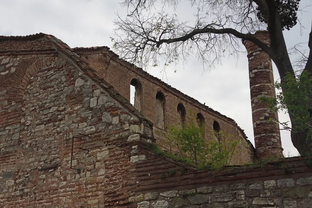 İmrahor İlyas Bey Camii'nin yan cephesi alttan; sıra hâlinde kemerli galeri pencereleri, kiremit kaplı duvar başlığı, sağda tuğla örgülü minare gövdesi ve çıplak ağaç.