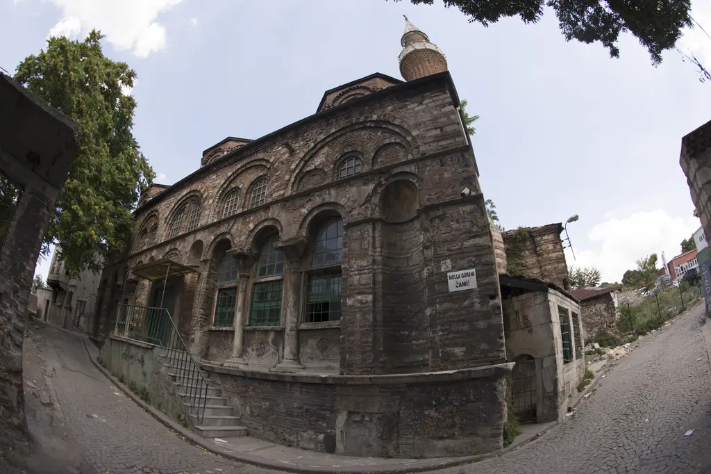 Vefa Kilise Camii'nin güneybatı köşesi geniş açıyla; yokuş parke sokak, revak sütunçeleri, yeşil tenteli merdiven, köşede cami levhası ve arkada minare.