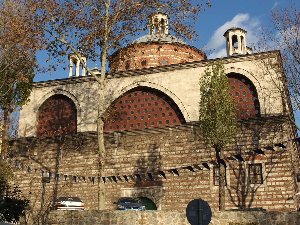 Tophane-i Âmire'nin yan cephesi; ağaçların arasından görünen kubbeler, kemerli duvar ve önünde yürüyen ziyaretçiler.
