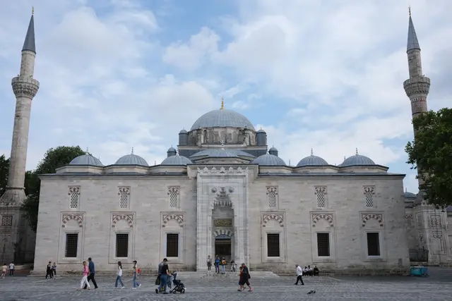 Beyazıt Camii önden; ortada kademelenen kubbeler, iki yanda birer ince minare, altta avlu duvarı ile mermer basamaklar ve meydanda yürüyen insanlar.
