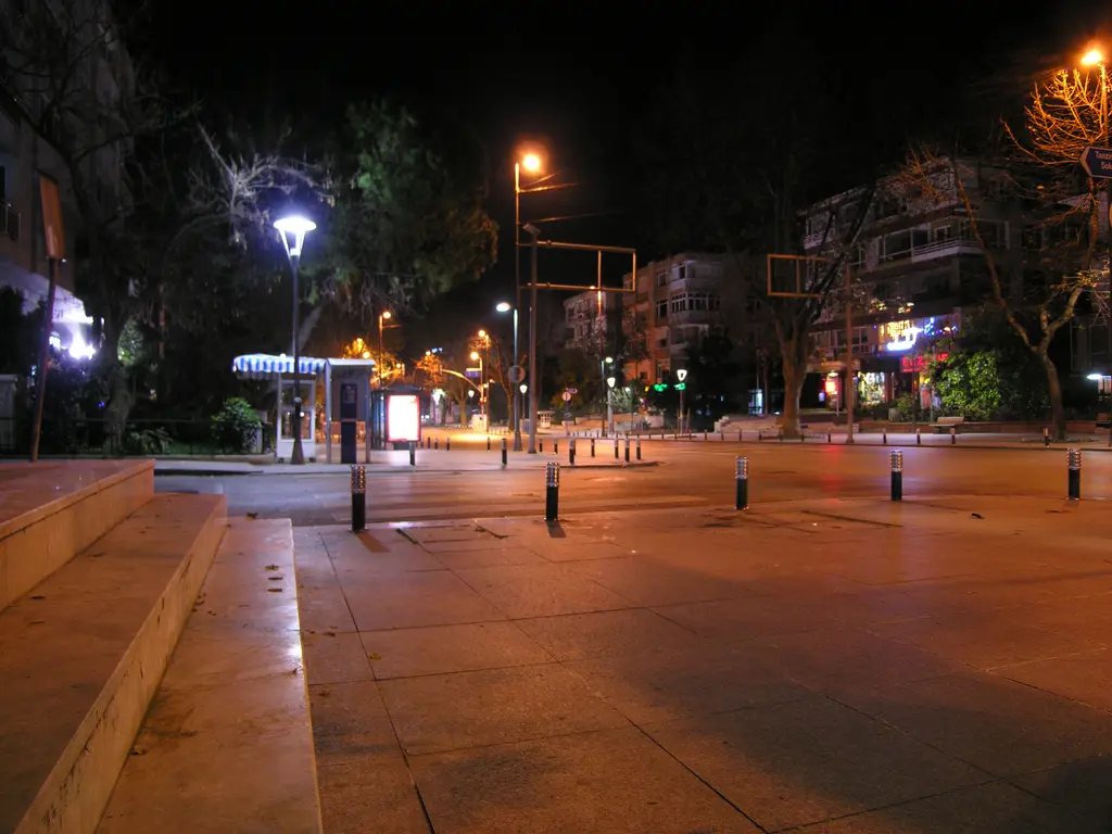 Gece Bağdat Caddesi'nde geniş kaldırım; turuncu sokak lambaları, bisiklet yolu, mavi ışıklı tabela ve ağaçlar.