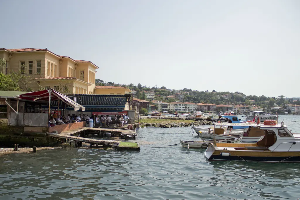 Çengelköy sahilinde demirli tekneler; solda sarı cepheli bina, suya uzanan teras ve uzakta Boğaz kıyısı.
