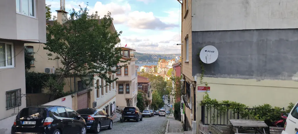 Kuzguncuk'ta Yakup Sokağı; iki yanda apartmanlar ve evler, park etmiş arabalar, ağaçlar ve uzakta Boğaz.
