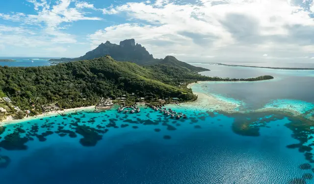 Bora Bora'nın havadan görünümü; Otemanu Dağı'nın yeşil yamaçları, mercan benekli turkuaz lagün, kıyıya dizilmiş su üstü bungalovlar ve parçalı bulutlu gökyüzü.
