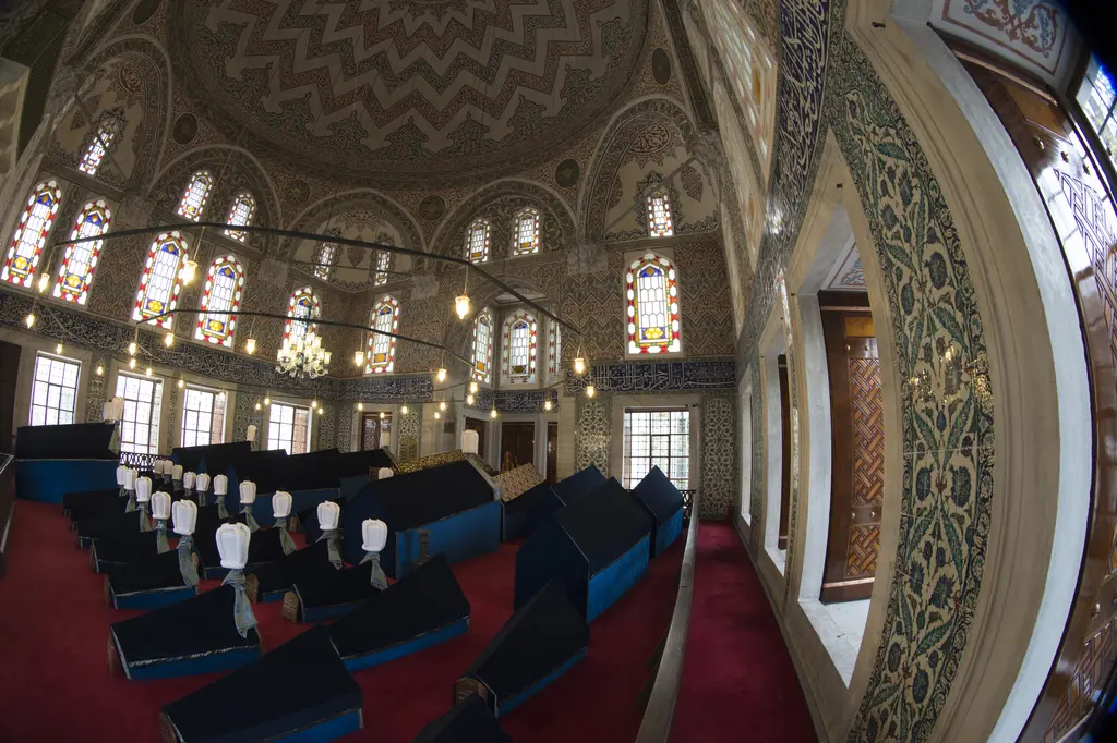 Sultan Ahmed Türbesi'ne geniş açılı bakış; kırmızı halı, koyu mavi örtülü sandukalar, beyaz sarıklar, çini kaplı duvarlar ve renkli camlı pencereler.