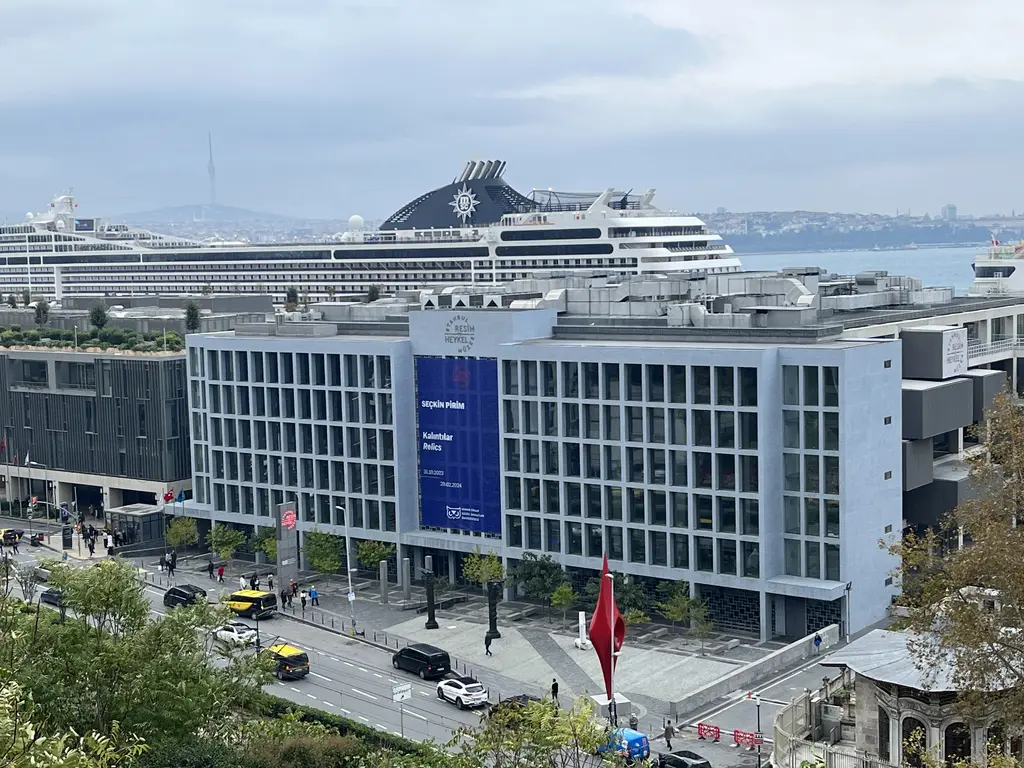 Galataport'ta mavi afişli cam cepheli bina ve arkasında yükselen kruvaziyer gemisinin üst katları; önde cadde ve ağaçlar.
