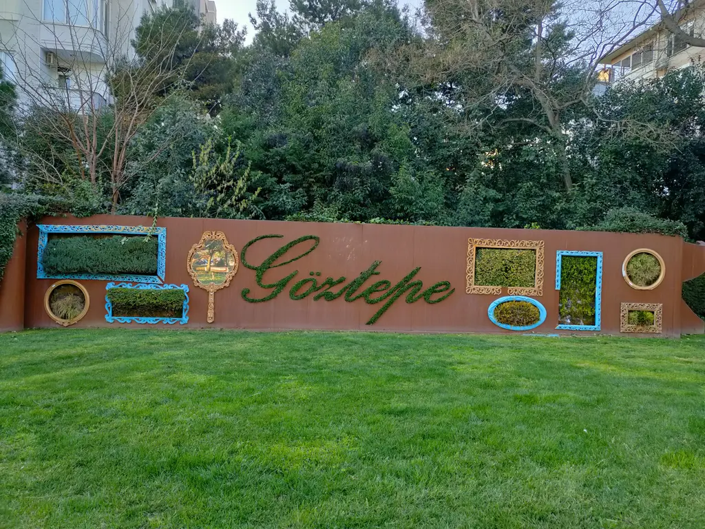 Göztepe Parkı'nda çimenlik; bitkilerle yazılmış Göztepe adı ve çerçeveli yeşil panolar taşıyan kahverengi duvar.