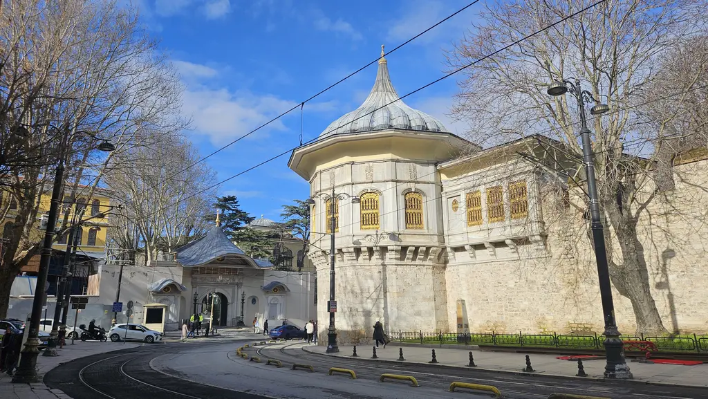 Alay Köşkü ve Alemdar Caddesi; burç üstünde soğan kubbeli köşk, sur boyunca uzanan kanat, solda Gülhane Parkı kapısı, tramvay rayları ve mavi gök.