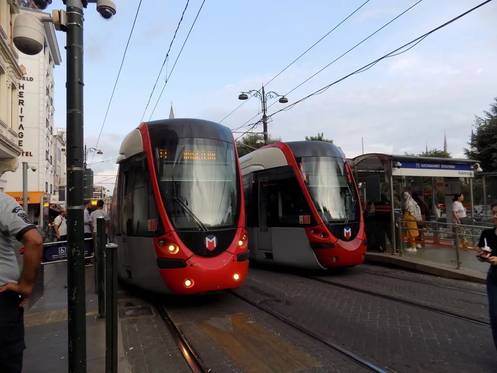 Sultanahmet'te durakta yan yana duran iki kırmızı beyaz tramvay; üstte elektrik telleri, perondaki yolcular ve arkada ağaçlar.