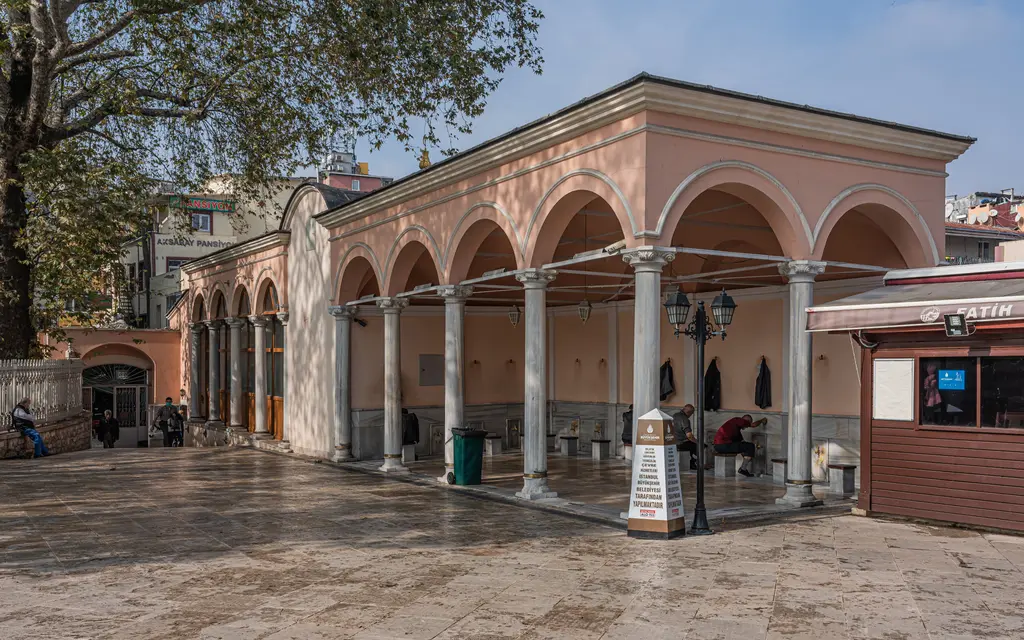 Pertevniyal Valide Sultan Camii'nin avlusunda kemerli revaklı yapı; ince sütunlar, pembe sıvalı duvarlar, ağaçlar ve ıslak taş zemin.