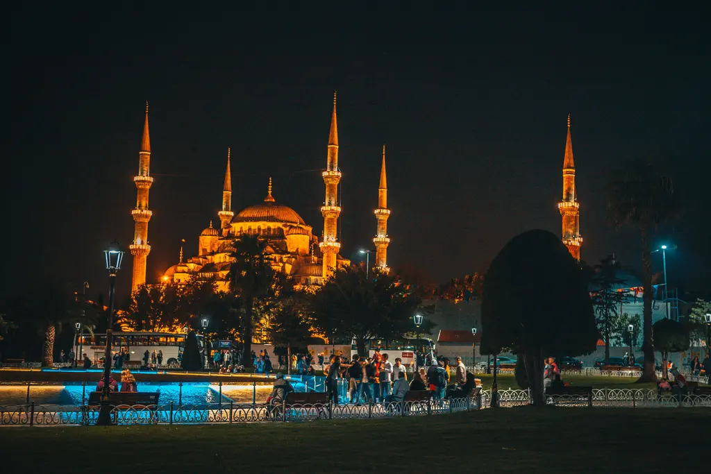 Gece Sultanahmet Camii; turuncu ışıkla aydınlatılan kubbeler ile altı minare, önde aydınlatılmış havuz ve çevresinde toplanmış kalabalık.