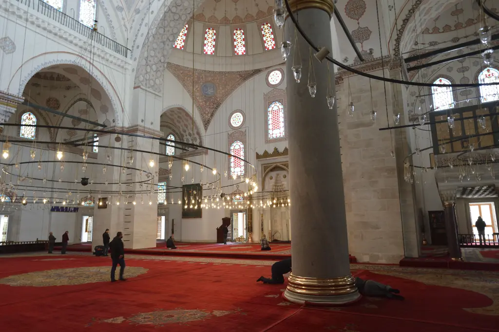Harim mihraba doğru; kırmızı halı, kalın granit sütun, halka biçimli alçak avizeler, arkada mermer mihrap ile minber ve namaz kılan insanlar.