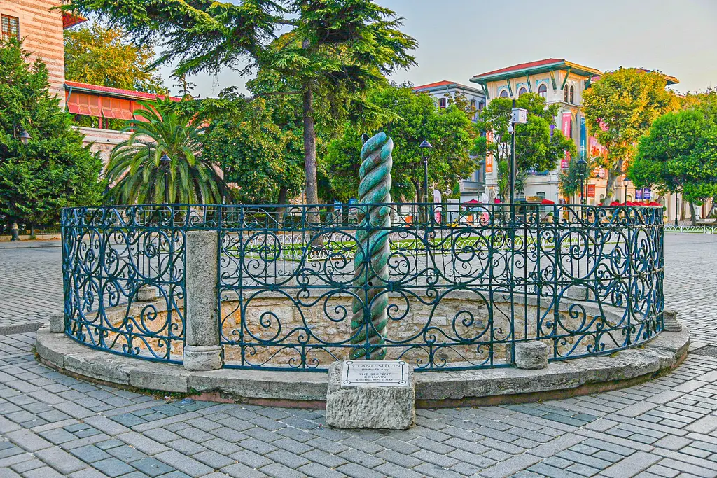 Yılanlı Sütun ve parmaklığı; önde taş döşeli meydan, arkada palmiyeler, ağaçlar ve Türk ve İslam Eserleri Müzesi'nin binası.
