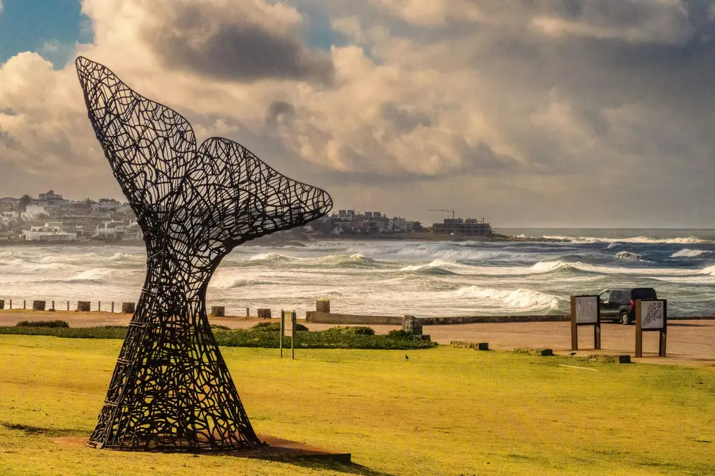 Uruguay'da Punta del Este kıyısında metal örgülü balina kuyruğu heykeli; arkada köpüklü dalgalar, kıyı evleri ve dramatik bulutlu gökyüzü.