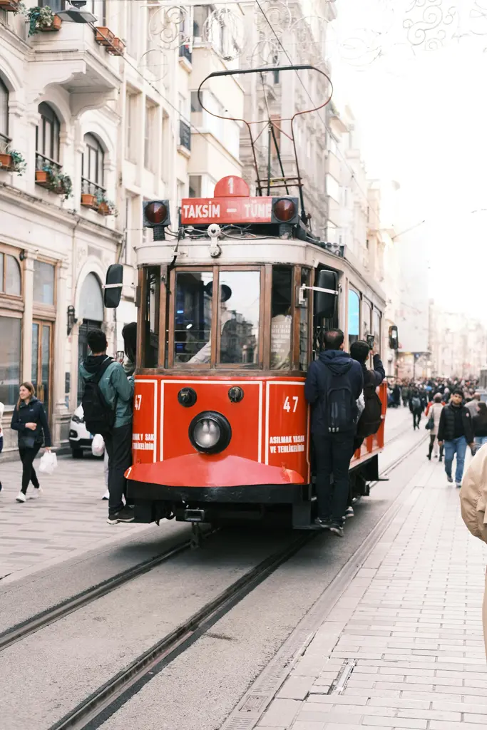 İstiklal Caddesi'nde kırmızı nostaljik tramvay; raylar üzerinde yaklaşan araç, iki yanda taş cepheli binalar ve arkada yürüyen insanlar.