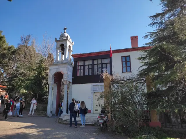 Büyükada'da Aya Yorgi Kilisesi; beyaz çan kulesi, ahşap çıkmalı manastır binası ve avlunun önünde gezinen ziyaretçiler.