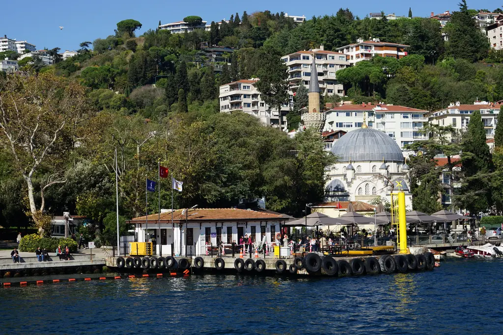 Bebek'te Boğaz'dan koy; kıyıda iskele binası, arkasında kubbeli ve tek minareli Bebek Camii, yamaçta ağaçlar ve evler.
