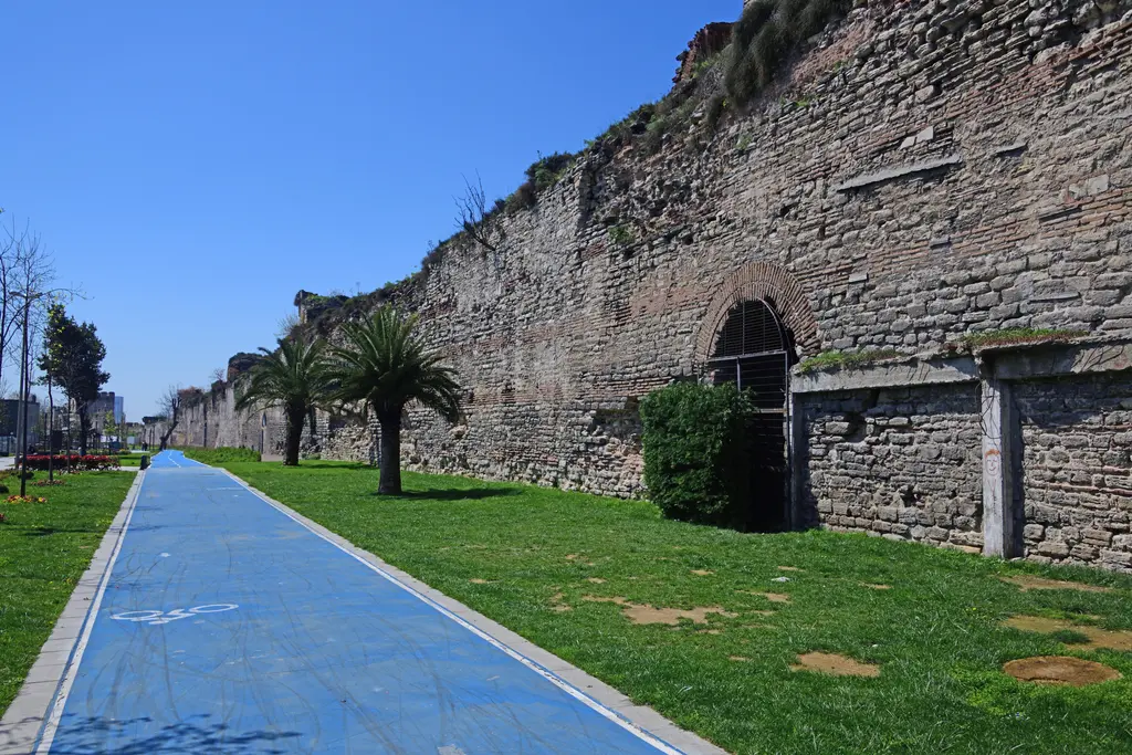 Sur boyunca uzanan yol; mavi boyalı bisiklet şeridi, kenarındaki palmiyeler ve çimenlik, sağda kemerli bir kapısı olan uzun duvar.