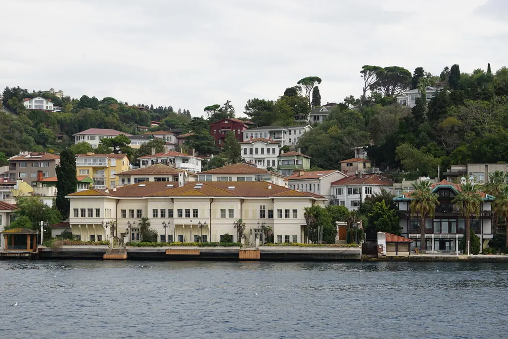 Saffet Paşa Yalısı ve çevresi; kıyıda uzun krem cepheli yalı, arkada yamaca dağılmış evler ve palmiyeler.