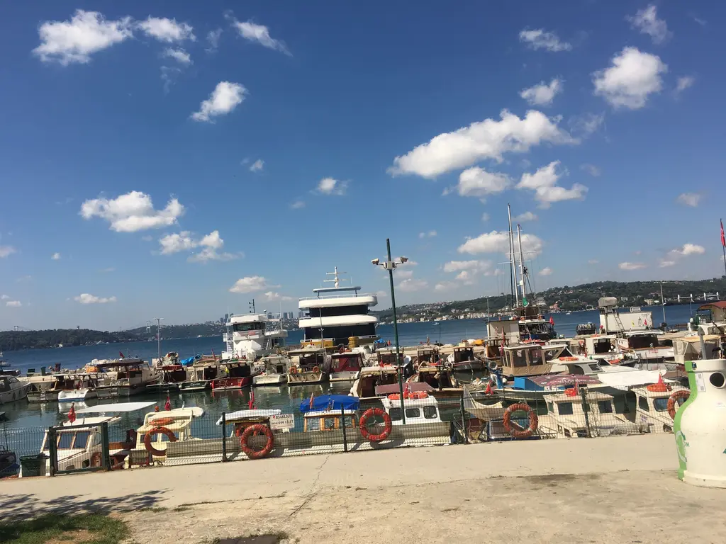 Üsküdar kıyısı; rıhtıma bağlı tekneler, arkada iskele yapıları ve parçalı bulutlu mavi gökyüzü.