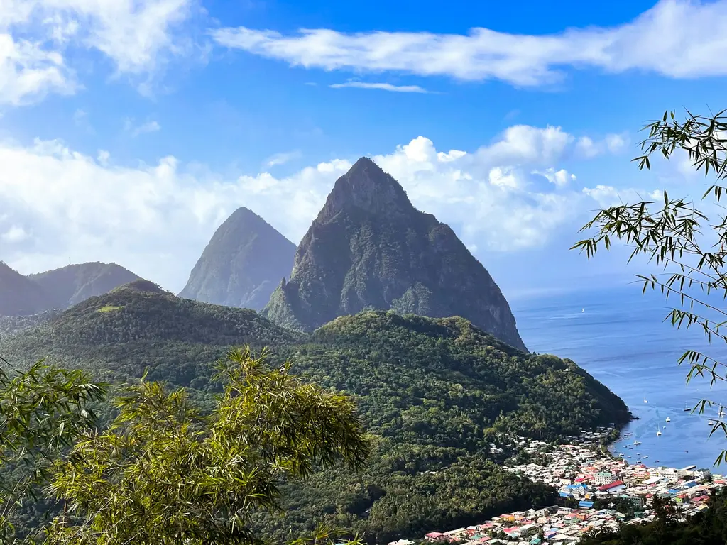 St. Lucia'da denizden dik yükselen ikiz Piton dağları; ormanla kaplı yamaçlar, altta Soufrière kasabasının renkli çatıları ve teknelerin demirlediği koy.