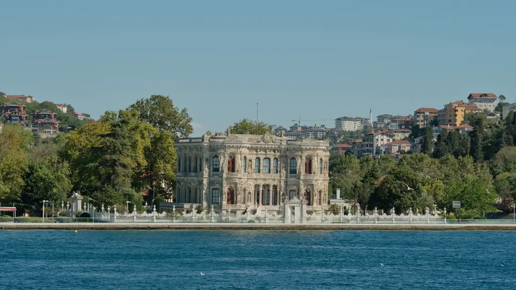 Boğaz'dan Küçüksu Kasrı; kıyıda yeşilliklerin arasında süslü kasır, önünde beyaz parmaklıklar, arkada evlerle dolu yamaç.