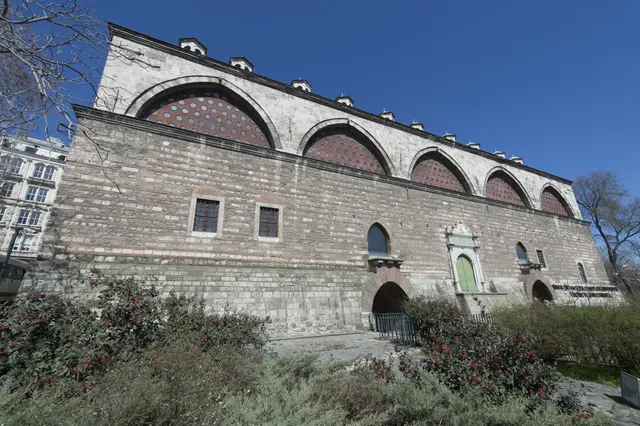 Tophane-i Âmire'nin uzun cephesi; kesme taş ve tuğla örgülü duvar, üstte sıra sıra kemerler, çatıda kubbe fenerleri ve mavi gökyüzü.