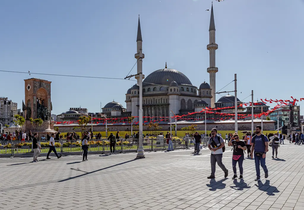 Taksim Meydanı'nda solda Cumhuriyet Anıtı, ortada iki minareli Taksim Camii; önde geniş taş zemin ve yürüyen insanlar.