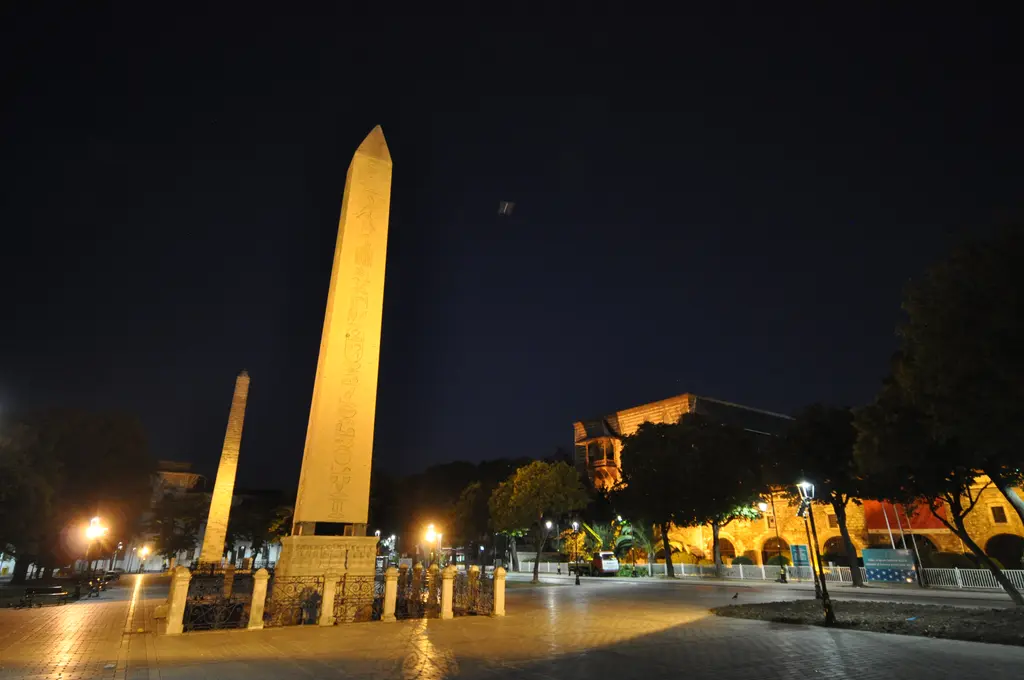 Gece Sultanahmet Meydanı; sarı ışıkla aydınlatılmış Dikilitaş, solda uzakta Örme Sütun, sokak lambaları ve karanlık ağaçlar.